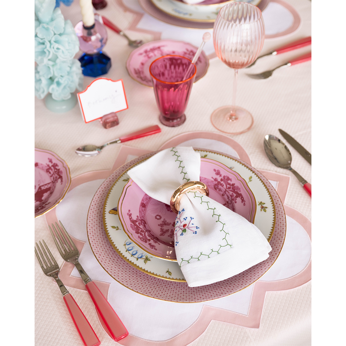 Pink scalloped-edge placemat with woven texture under floral patterned dinnerware on a table setting.