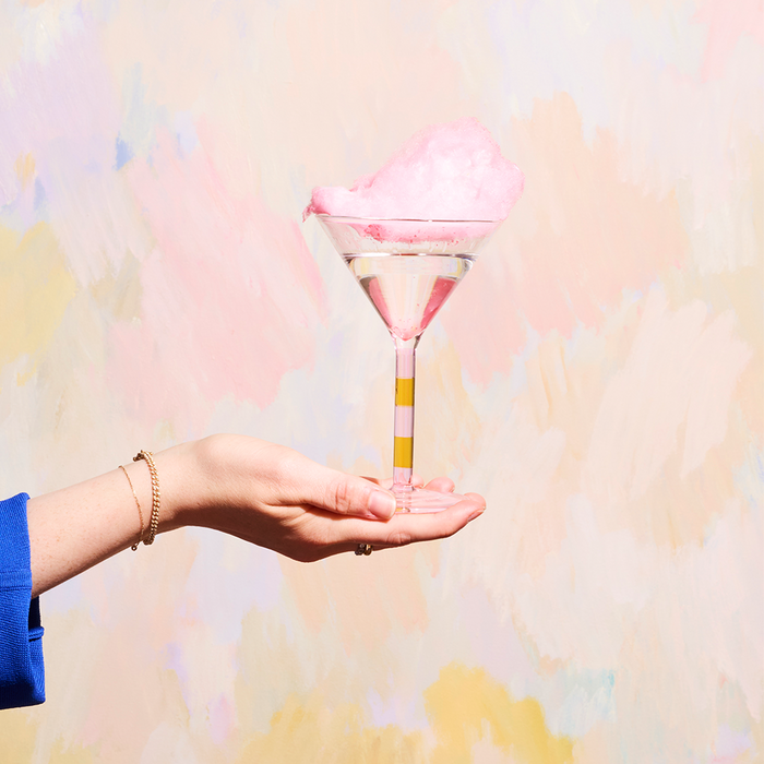 Clear martini glass with alternating pink and amber striped stem, filled with pink crushed ice, held by hand against pastel.