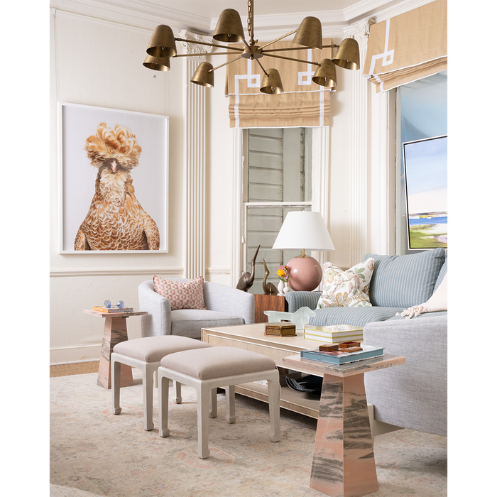 A styled living room interior featuring a large framed art print of a rooster on the wall above a light gray armchair. The room includes a light blue sofa, a wooden coffee table, a chandelier, and a window with beige blinds.